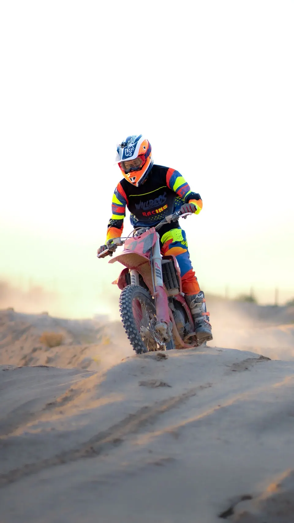 A motocross rider in bright orange and blue gear riding a dirt bike over sand